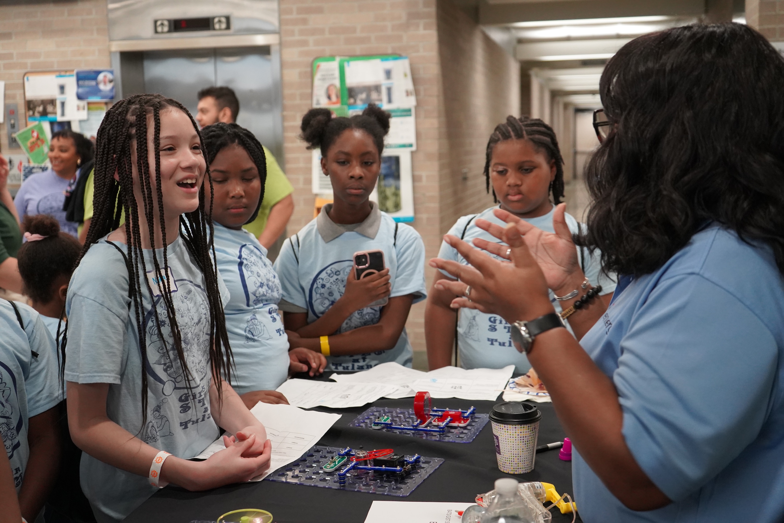 Young girls in light blue shirts listen to a woman at a science fair table.