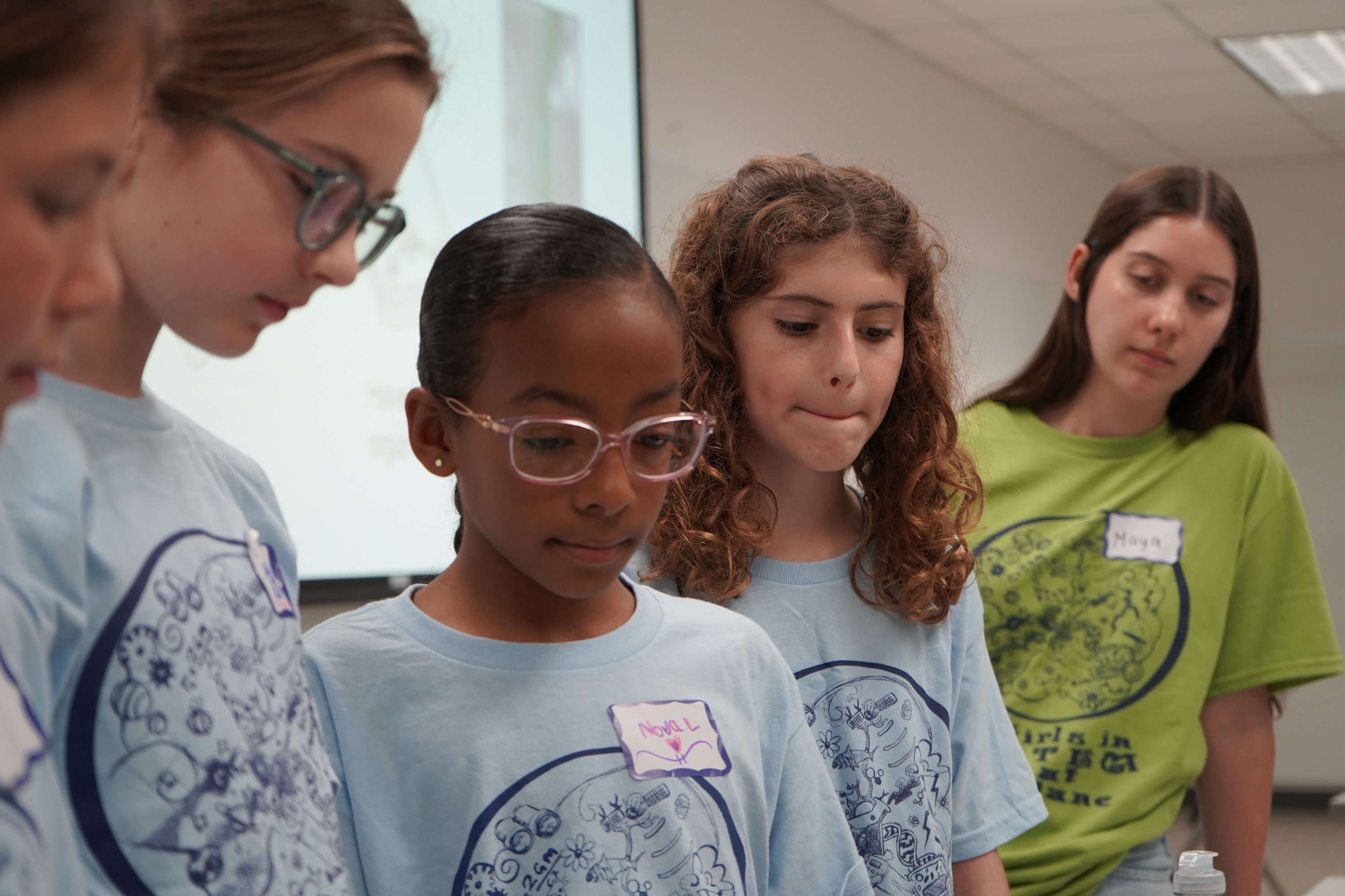 Group of diverse girls in light blue t-shirts looking down intently.