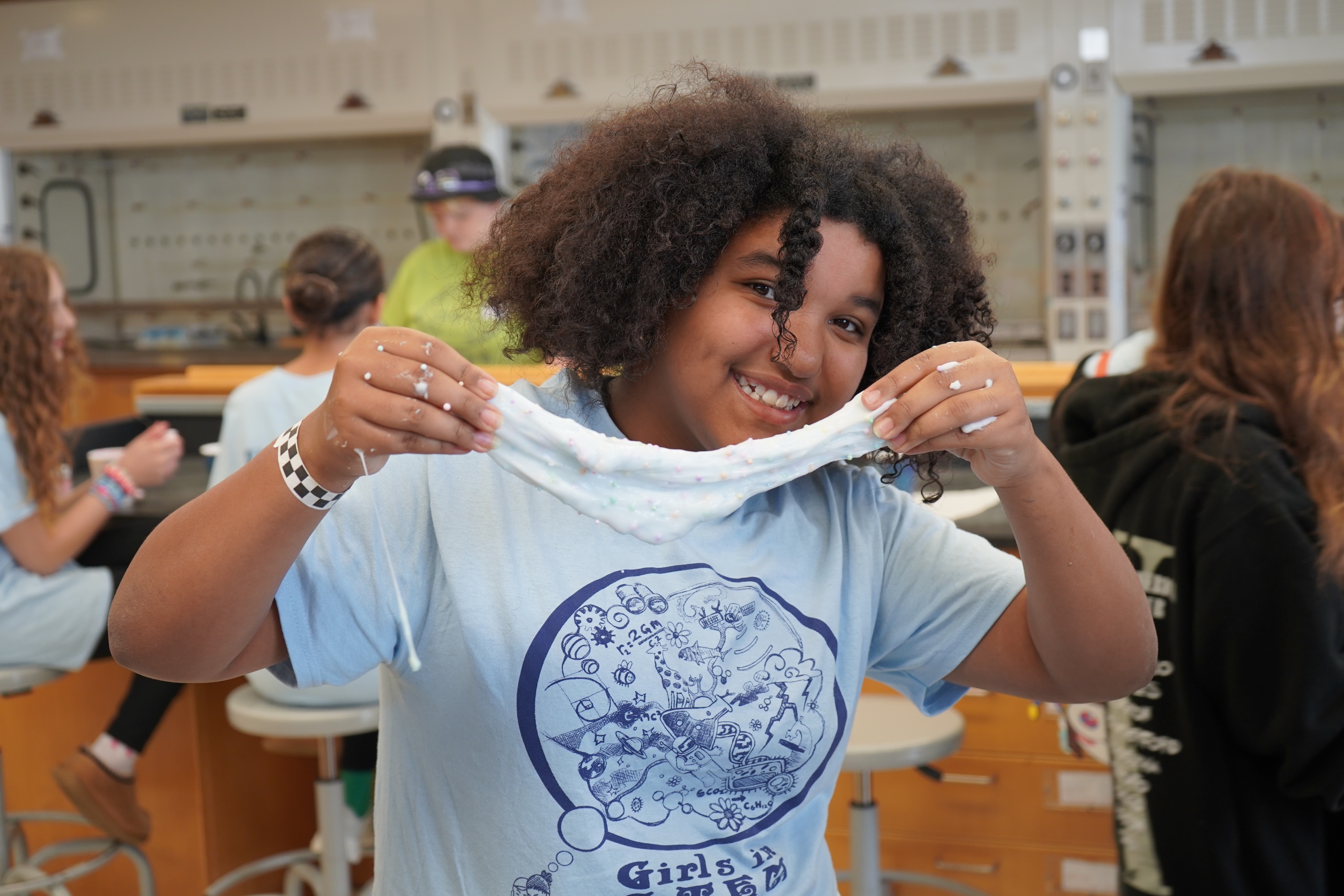 A smiling girl with curly hair holds up a string of white slime in a classroom.