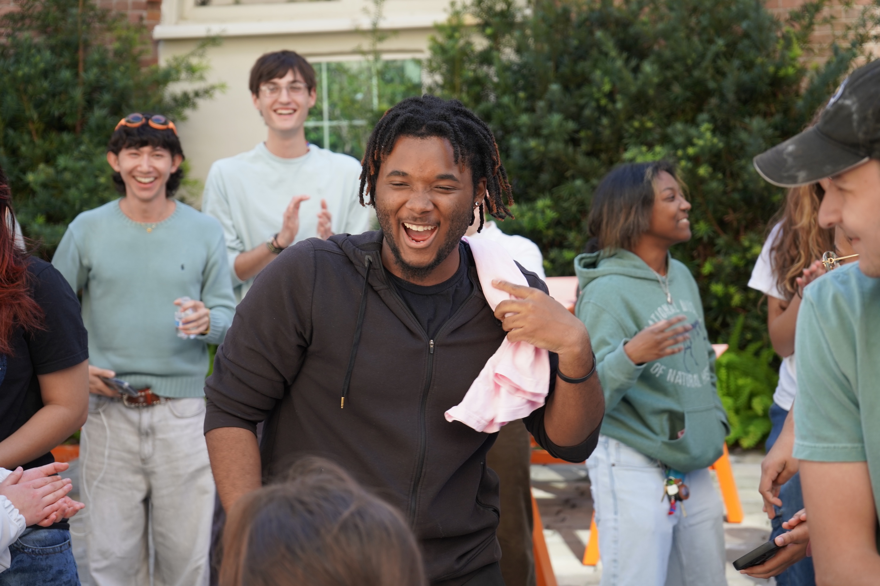 A Black man with dreadlocks laughs, holding a pink cloth, surrounded by a diverse group of friends.