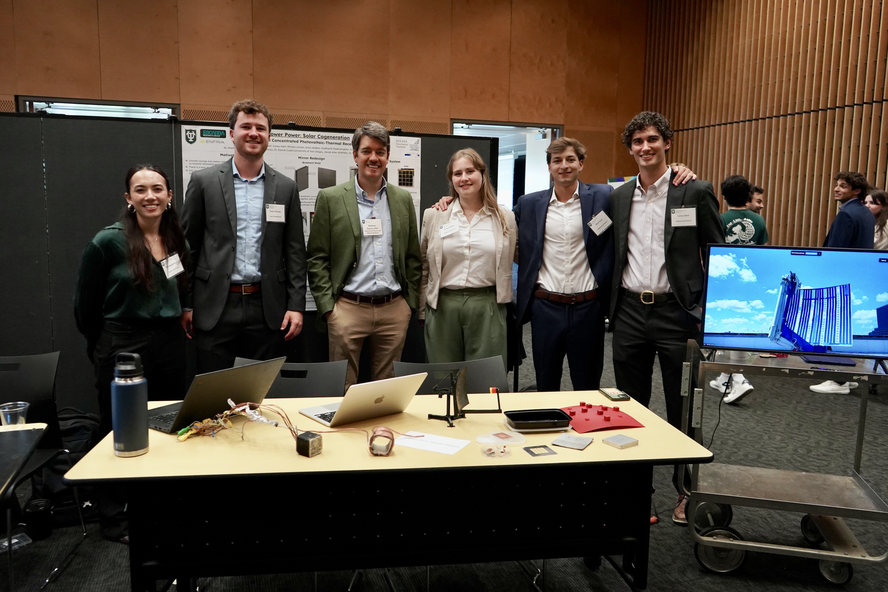 Six smiling people stand behind a table with laptops and a monitor displaying a graphic.