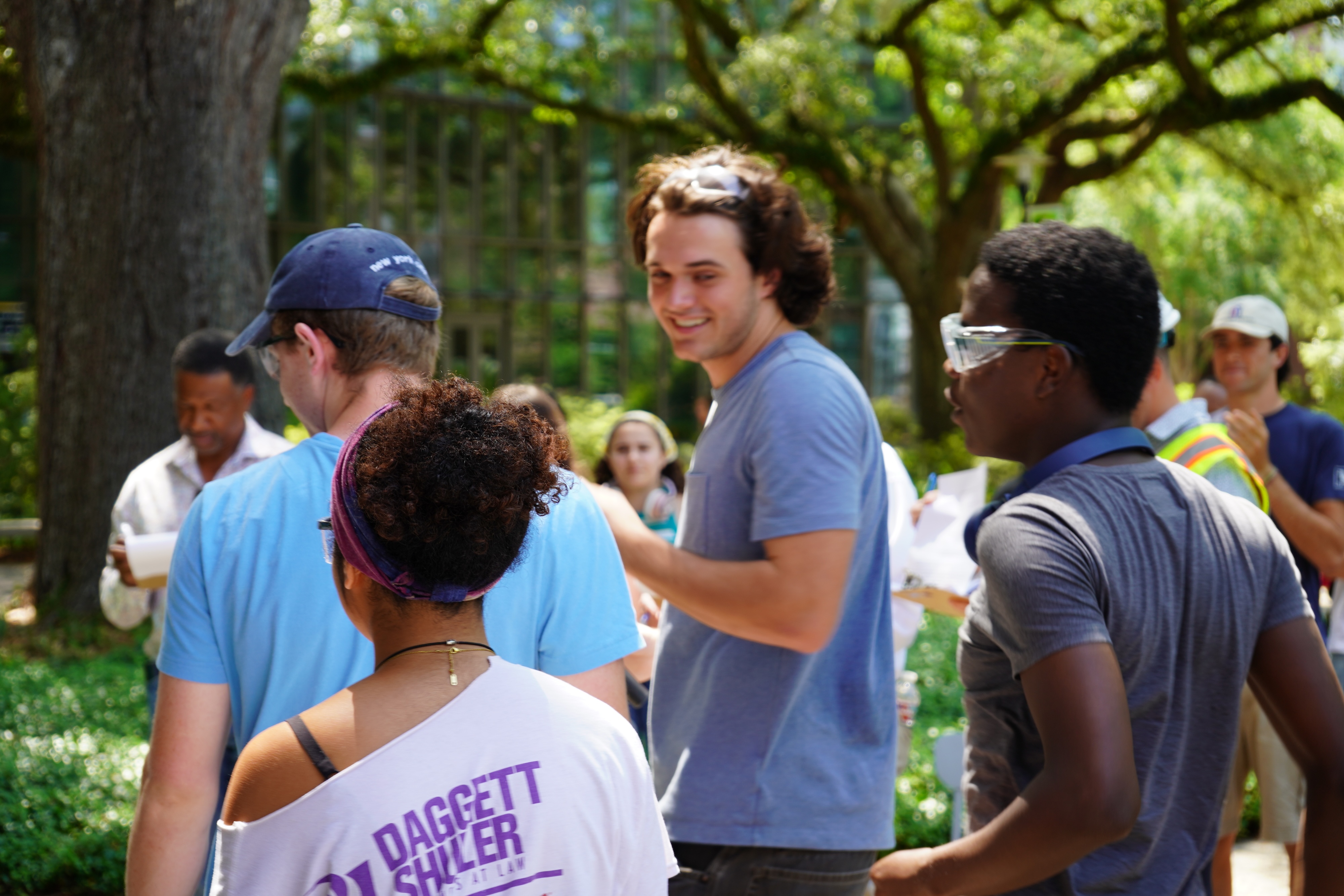 Smiling young man in blue shirt and sunglasses talks to a group of people outdoors.