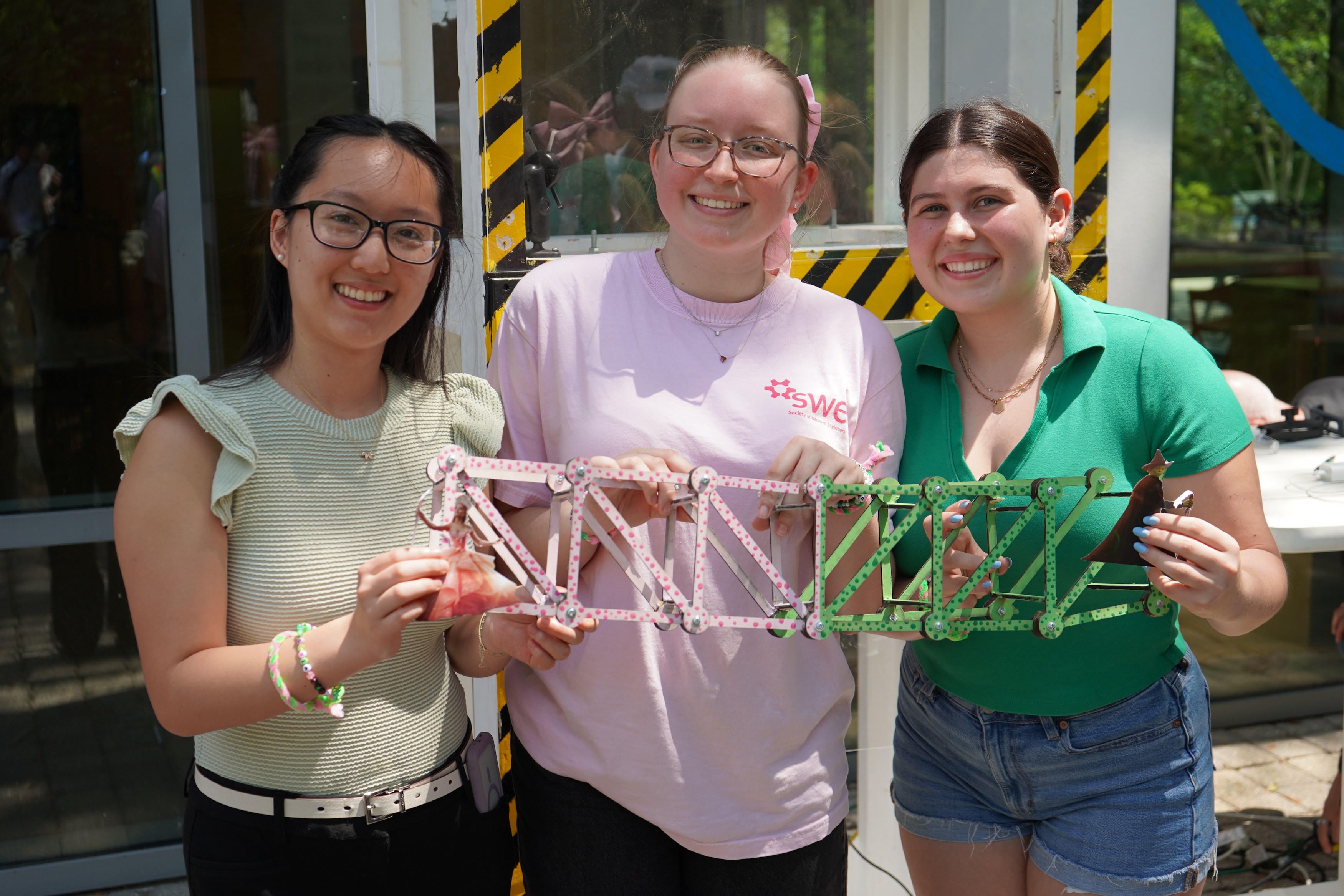 Three smiling young women hold a colorful, intricate model bridge.