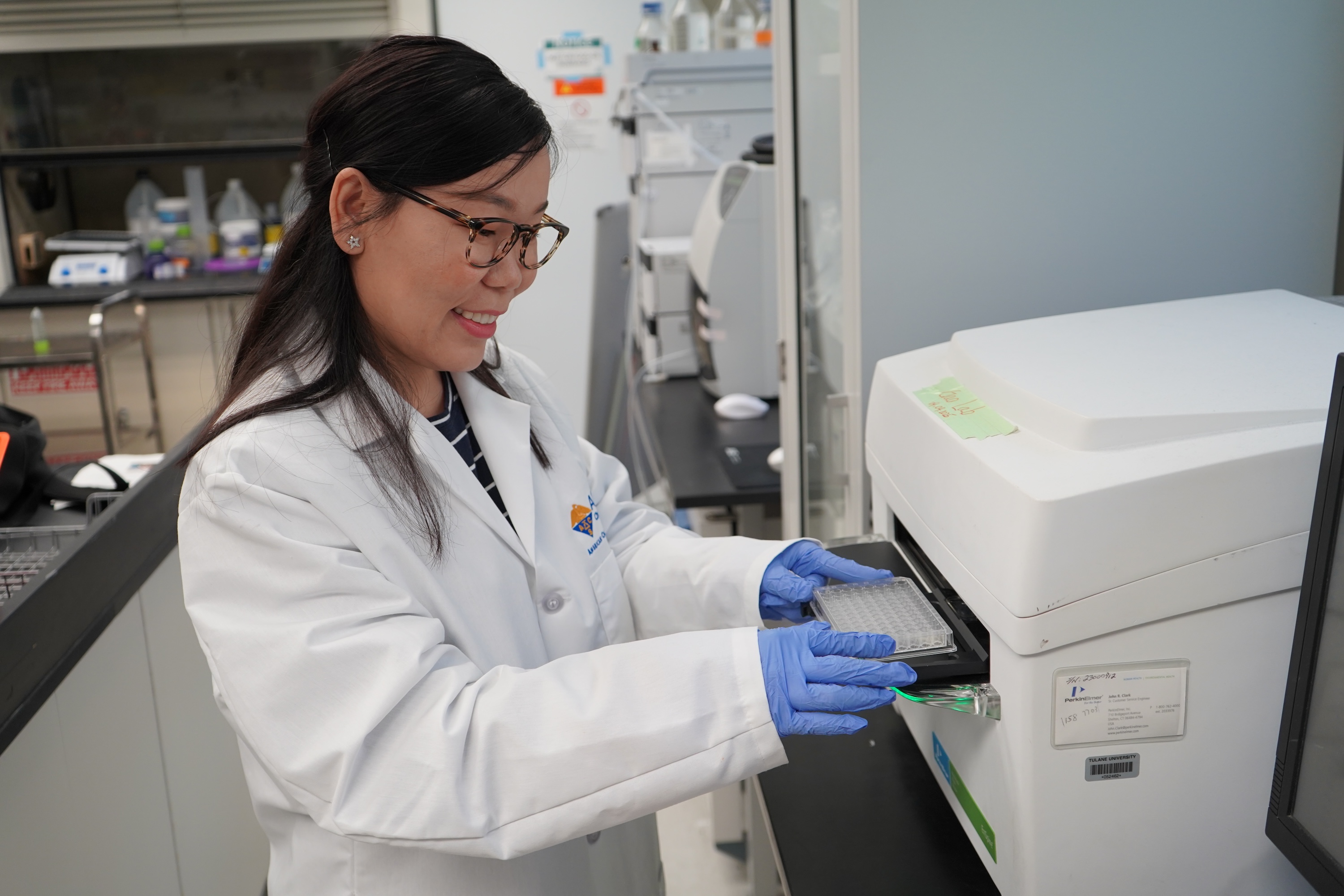 Woman in lab coat and gloves inserts a tray into a machine.