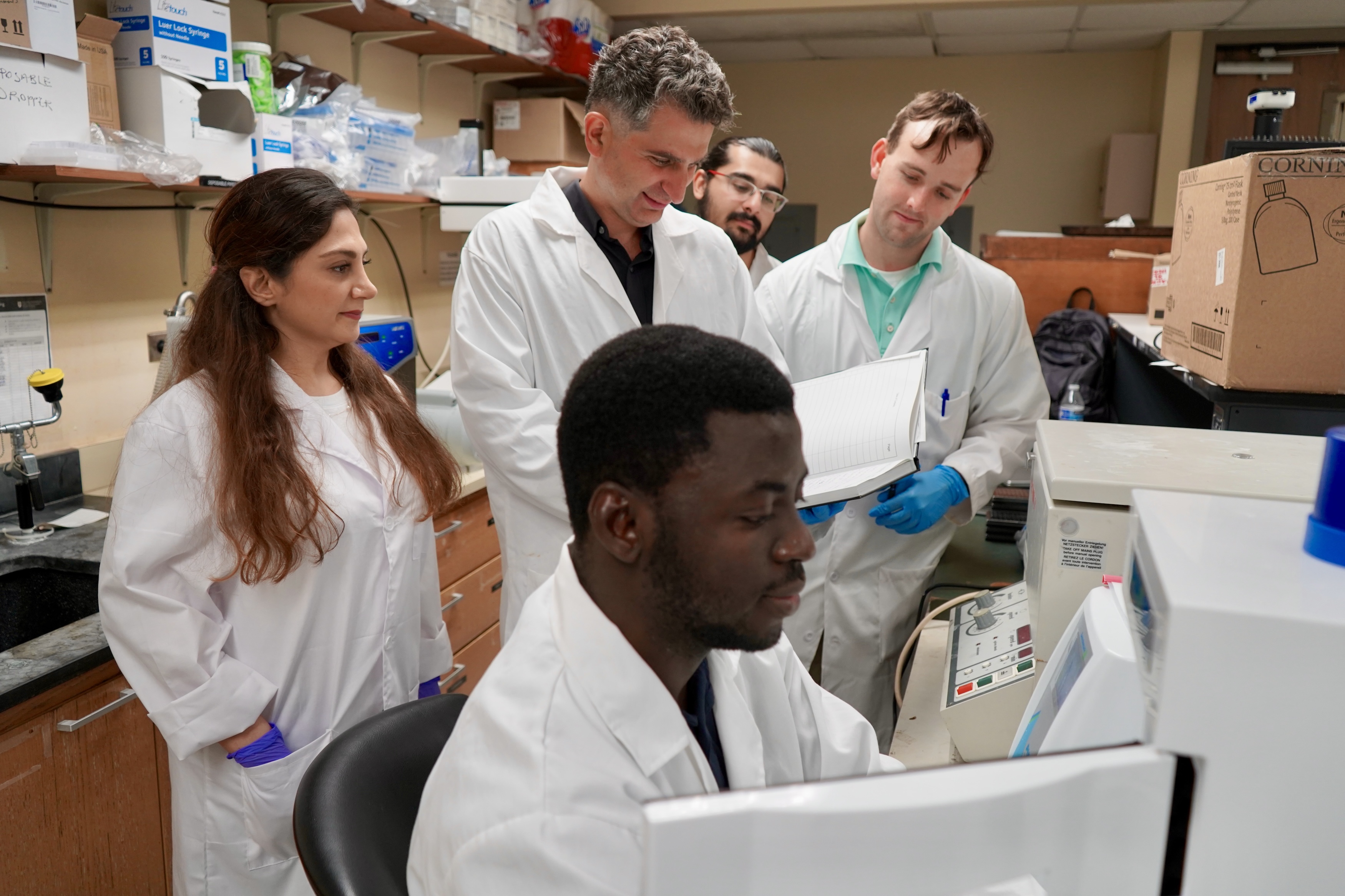 Scientists in lab coats gather around a computer in a lab.