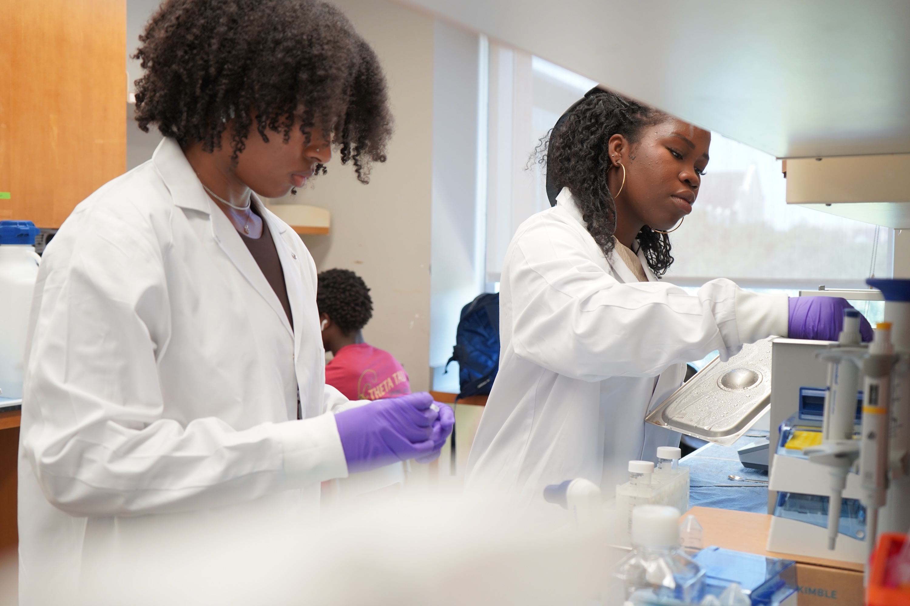 Two women in lab coats work at benches, wearing purple gloves.