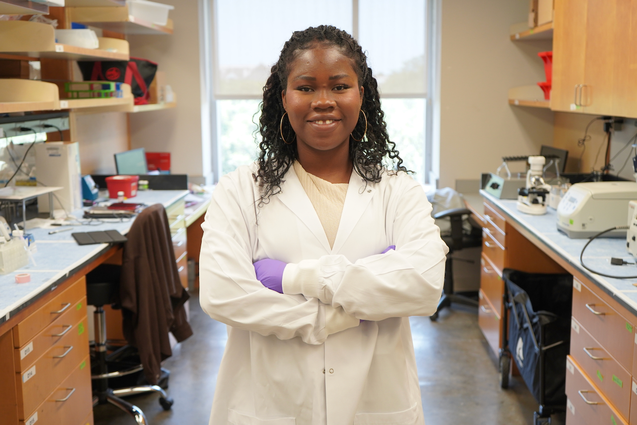 A woman in a lab coat and purple gloves crosses her arms, smiling.