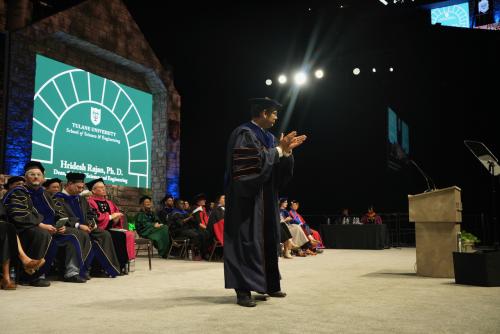 A man in academic regalia claps as a large screen displays university logos.