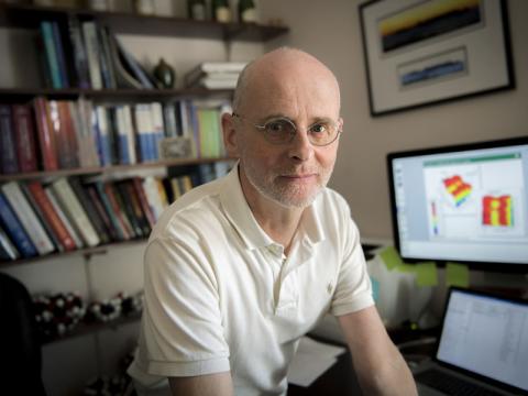 Bald man in glasses wearing a polo shirt at a desk with bookshelves and computer monitors.
