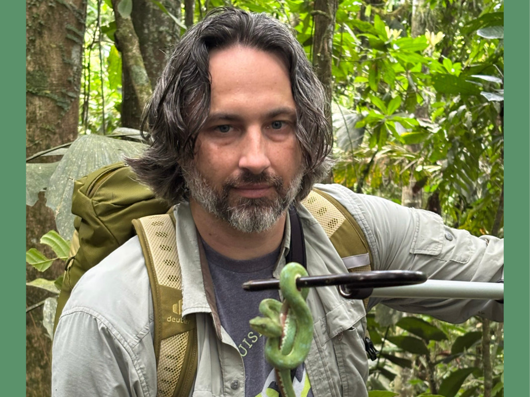 Man wearing a backpack and light colored shirt holds a green snake.