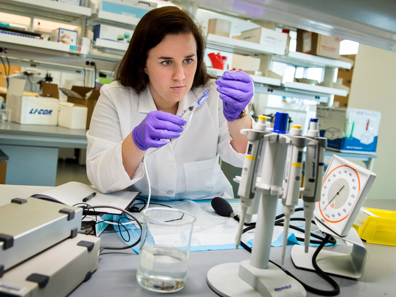 Woman in a lab coat with purple gloves holds a pipette over a beaker.