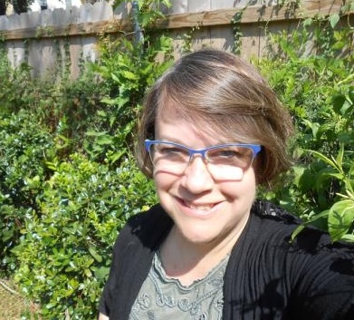A woman with short brown hair and glasses smiles at the camera in front of lush green foliage.