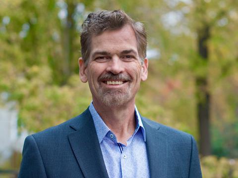 Man in a blue shirt and suit jacket smiles outdoors.