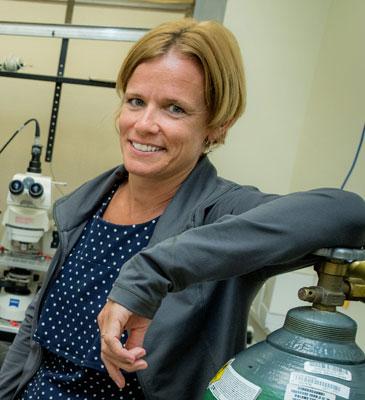 Woman smiling in a lab next to a gas cylinder and microscope.