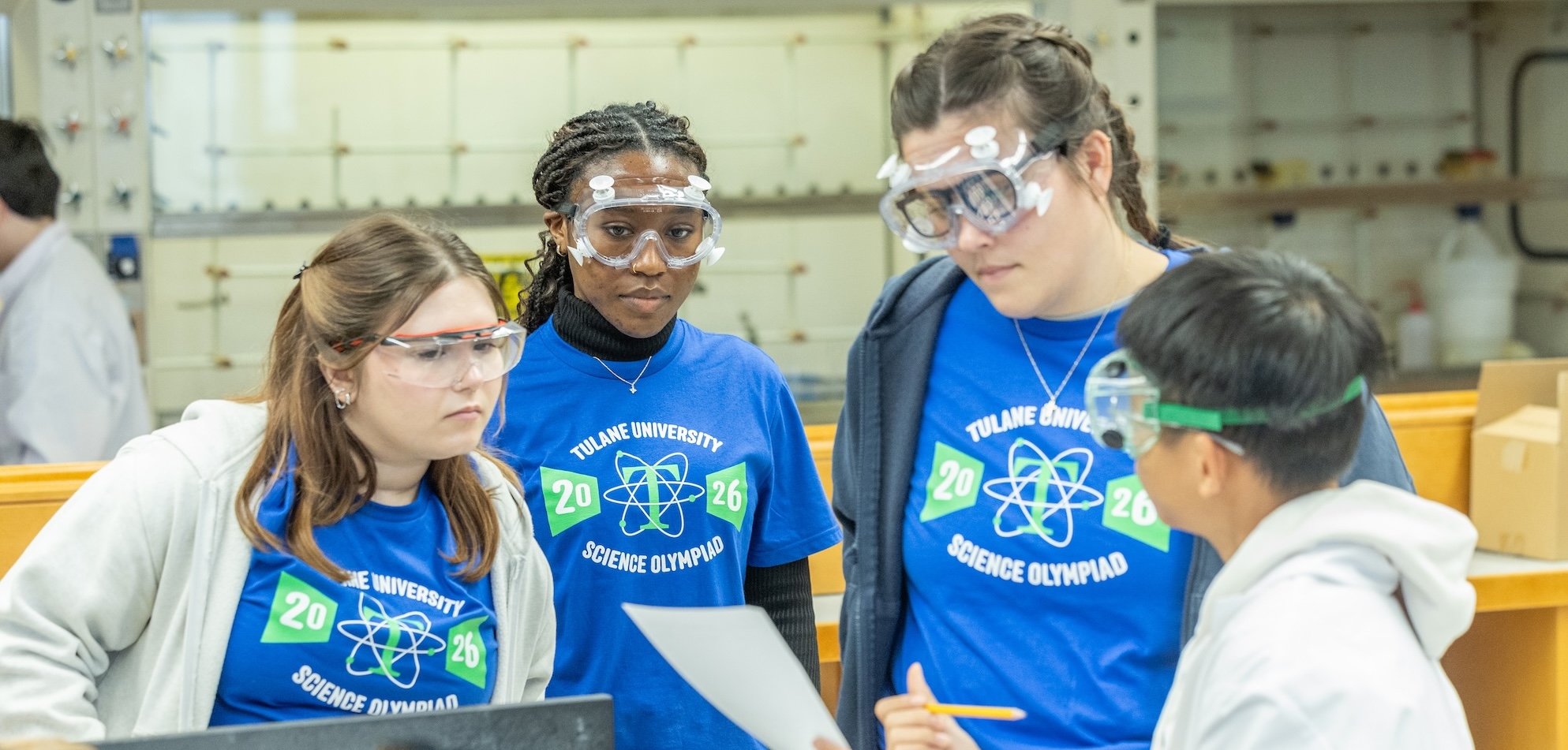 Four students in safety goggles look at a document in a lab.