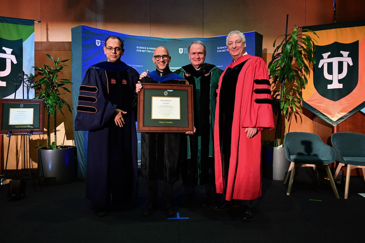 Four men in academic regalia stand on a stage holding a framed award.