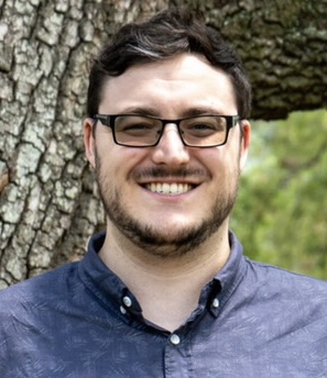 A smiling man with glasses and a blue shirt stands in front of a tree.