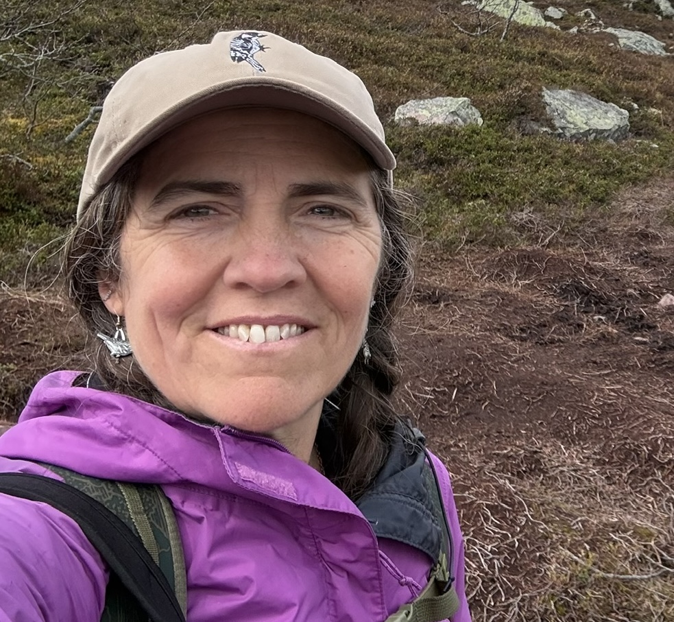 Woman wearing a purple jacket and tan baseball cap smiles outdoors on a rocky, grassy hillside.