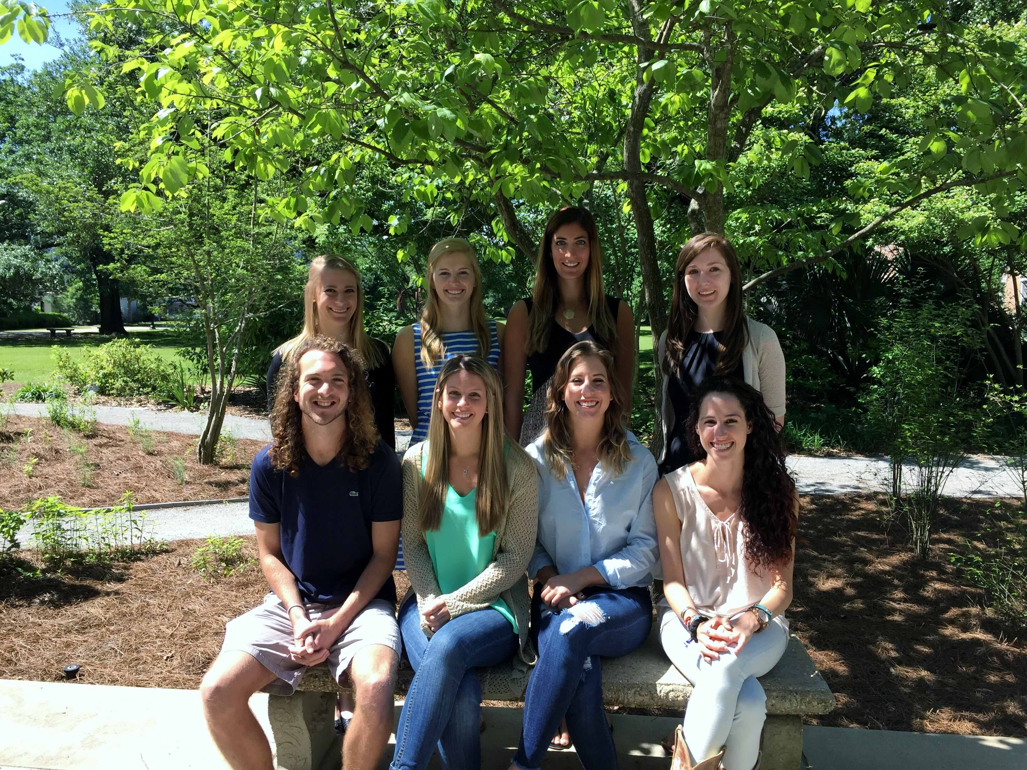 Photograph of a group of eight young adults smiling outdoors under a tree.