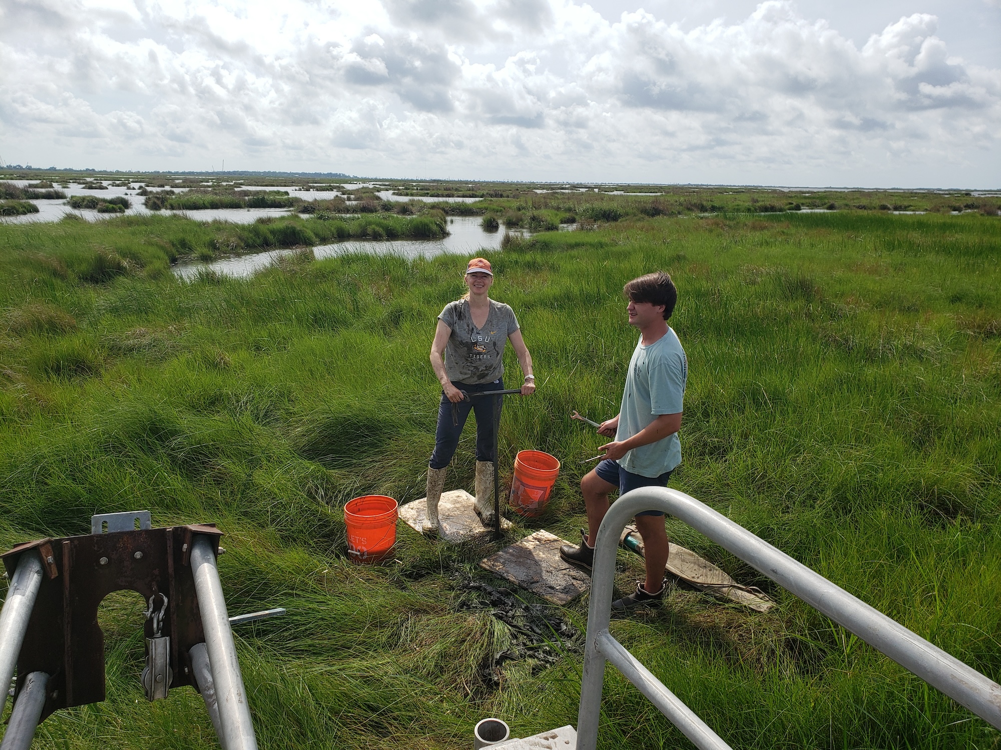 Photograph of two people standing in a grassy wetland with orange buckets.