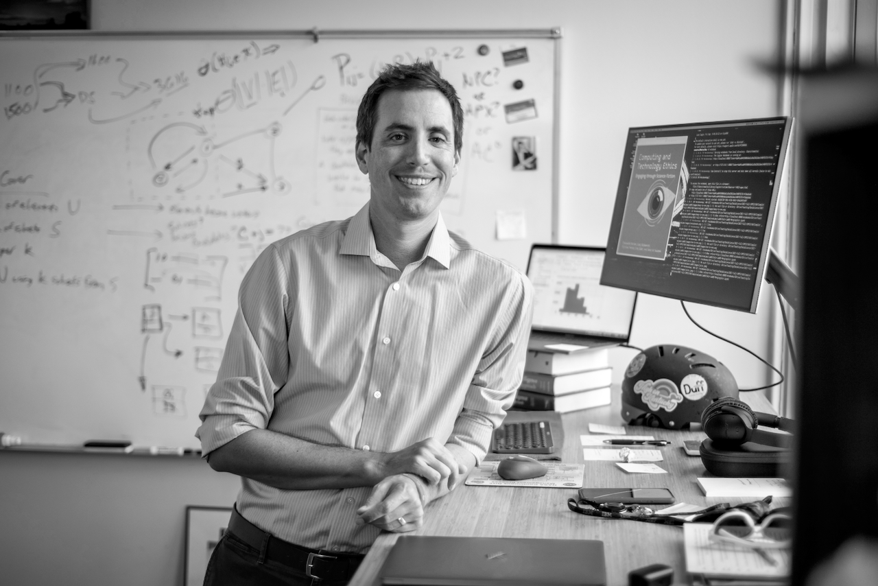 Black and white photo of a man leaning on a desk in an office, with a whiteboard behind him.