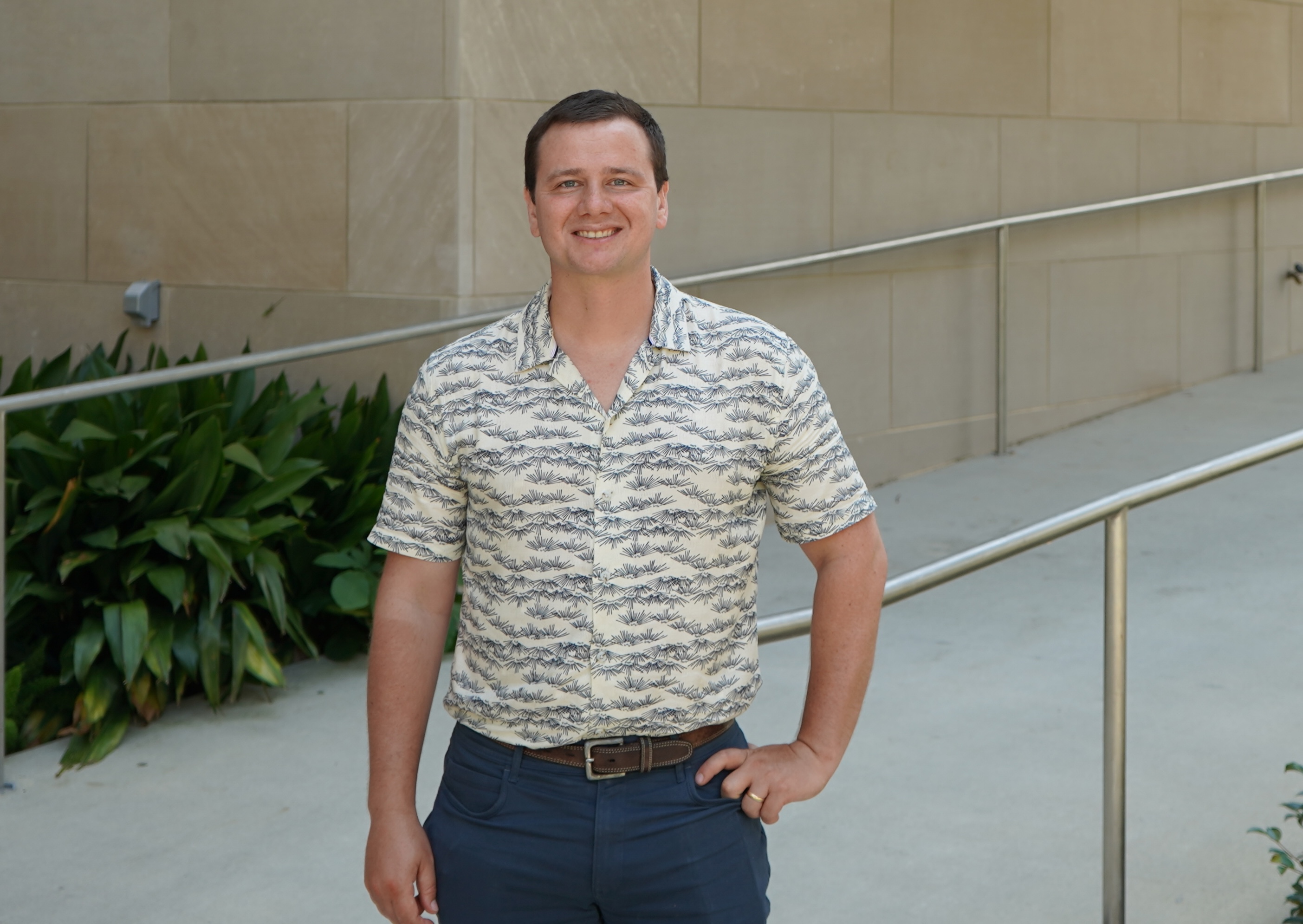 Photo of a smiling man wearing a patterned shirt standing outside near a building.