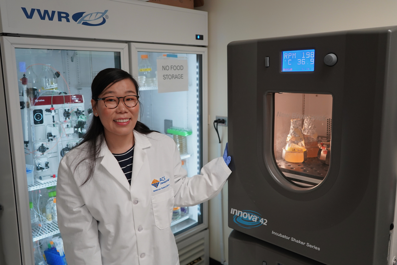 Scientist in lab coat next to a lab incubator holding flasks.