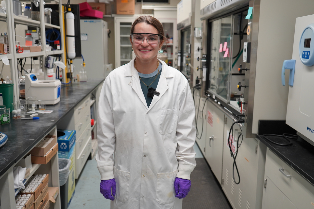Young woman in lab coat, goggles, and gloves smiles in a laboratory.
