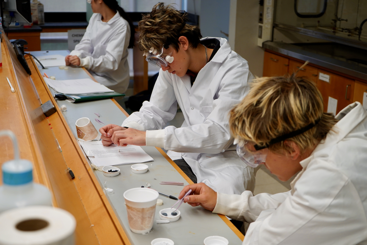Students in lab coats and goggles work at a science bench.