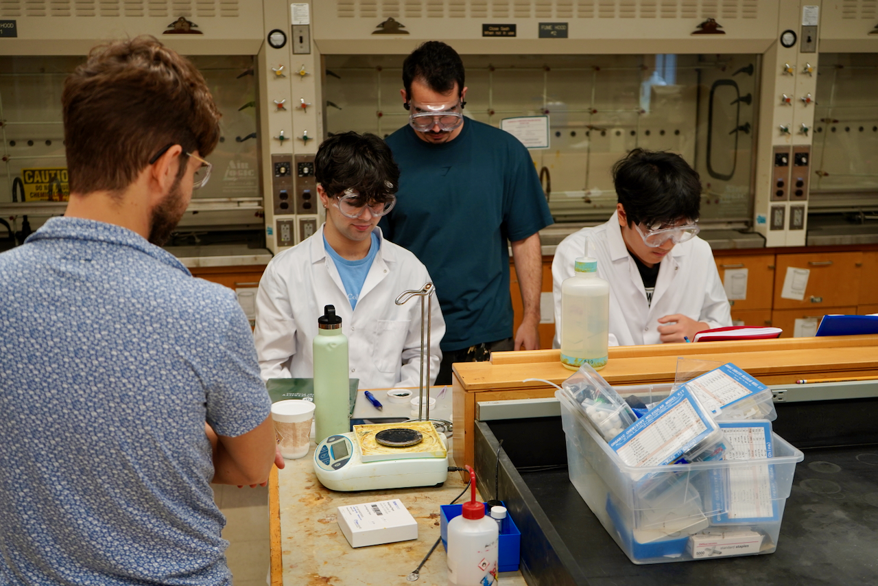 Four students wearing safety goggles work together in a chemistry lab.