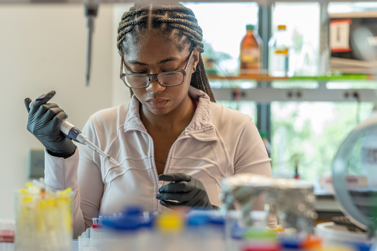 A Black female scientist wearing glasses and black gloves uses a pipette in a laboratory.