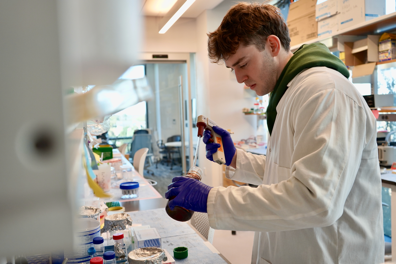 Male scientist in white lab coat pours liquid from a flask.