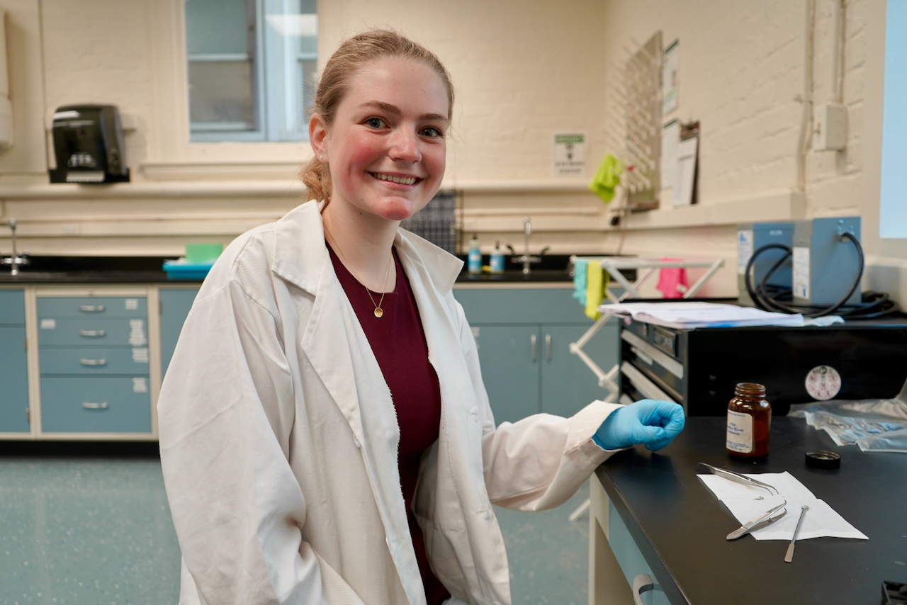 Girl in white lab coat and blue gloves at a lab desk smiling
