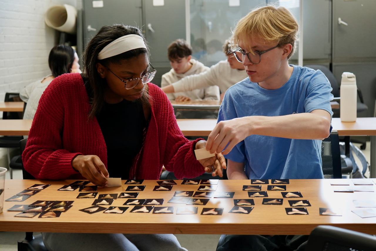 Two students play a card game at a table, with others in the background.