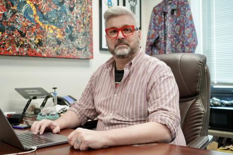 A man with red glasses and gray hair sitting at a desk with a laptop.