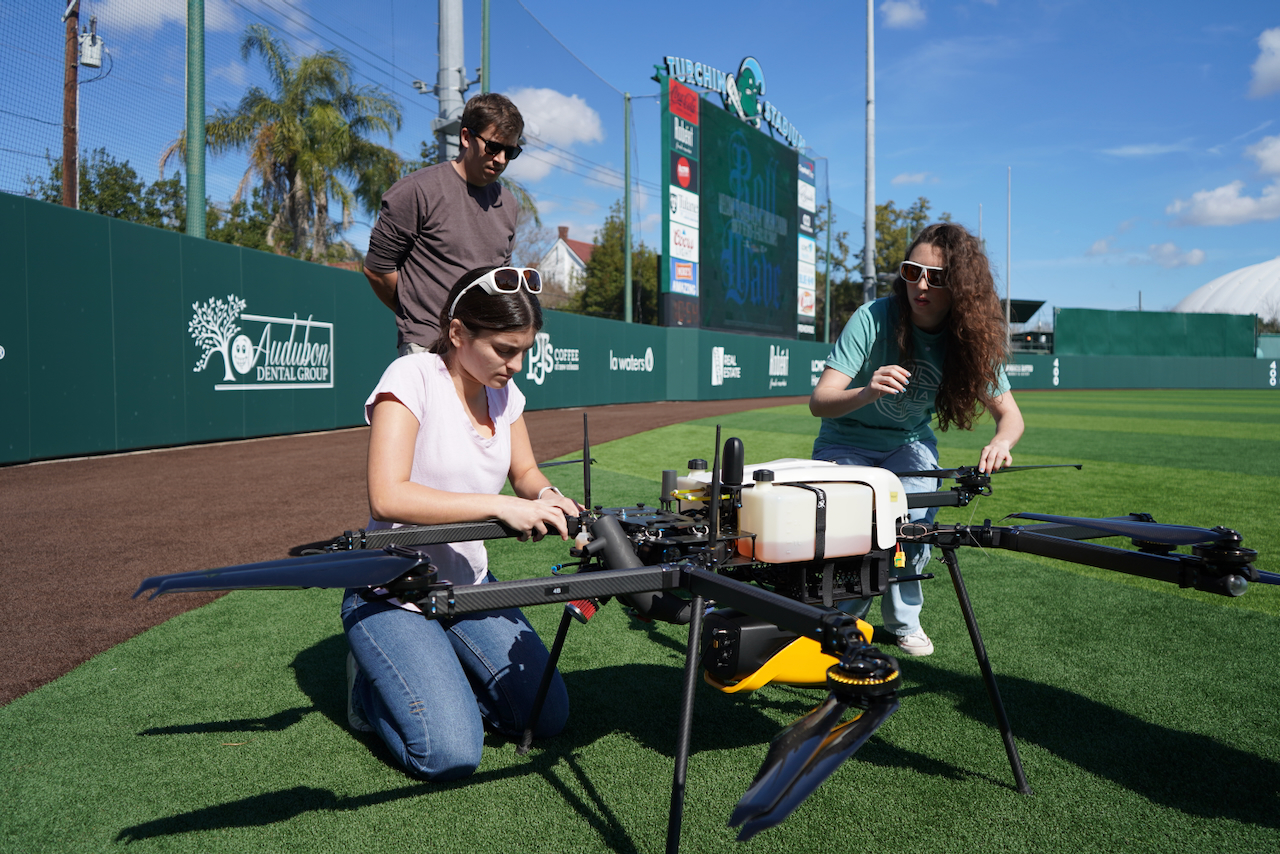 Photograph of three people preparing a large agricultural drone on a baseball field.
