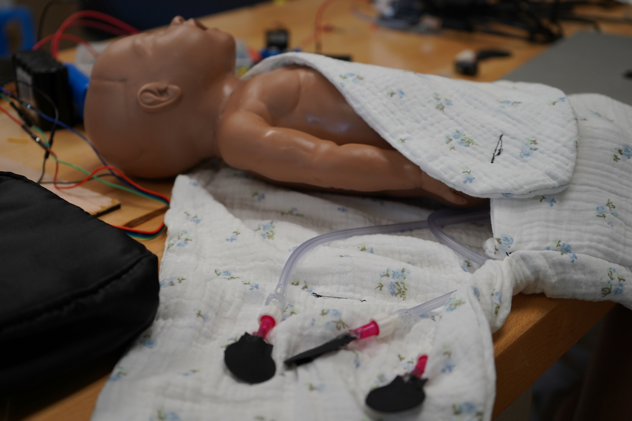 A baby mannequin in a white blanket with tubes attached on a wooden table.
