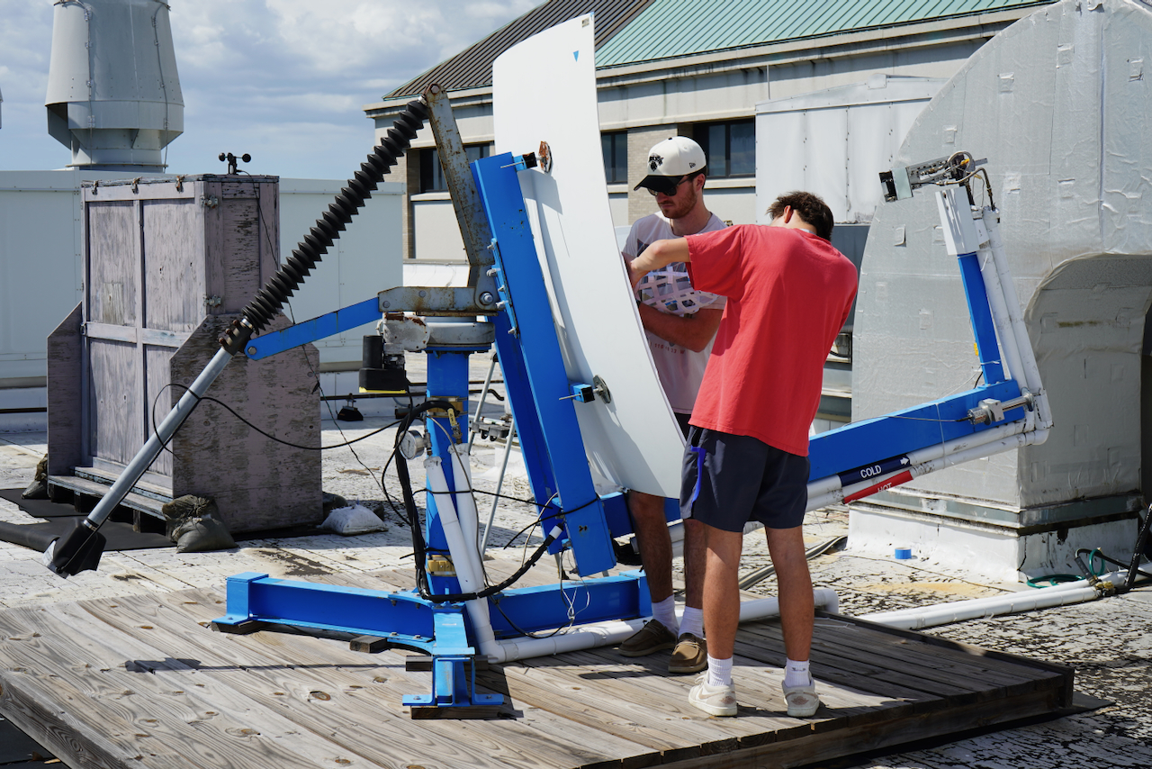 Photograph of two men adjusting a large, curved solar panel on a blue metal frame on a rooftop.