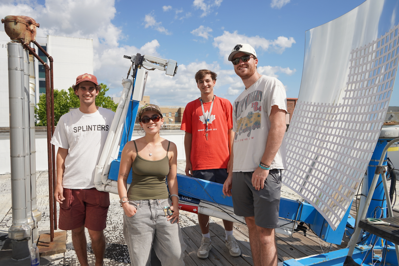 Four people stand next to a large parabolic dish on a rooftop.