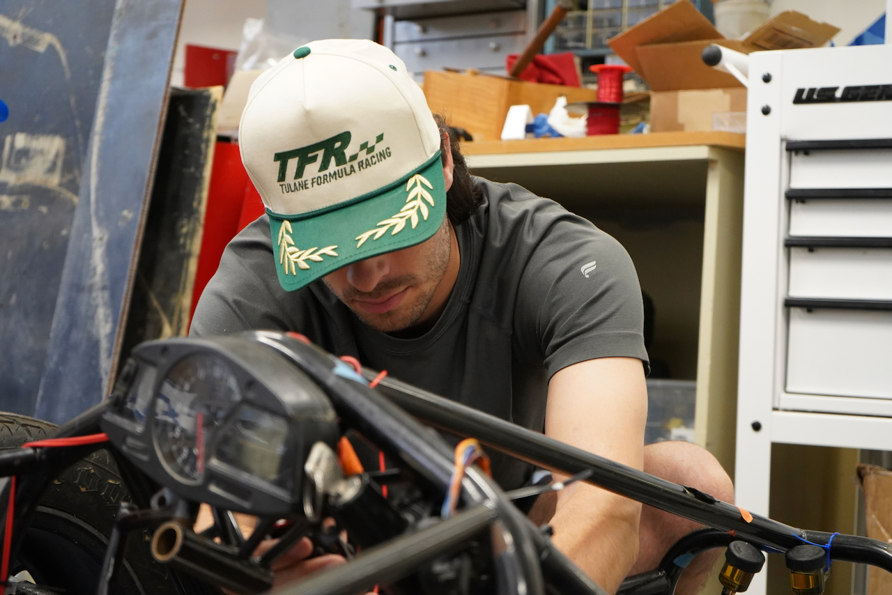 Photograph of a man wearing a TFR cap working on a race car frame in a cluttered garage.
