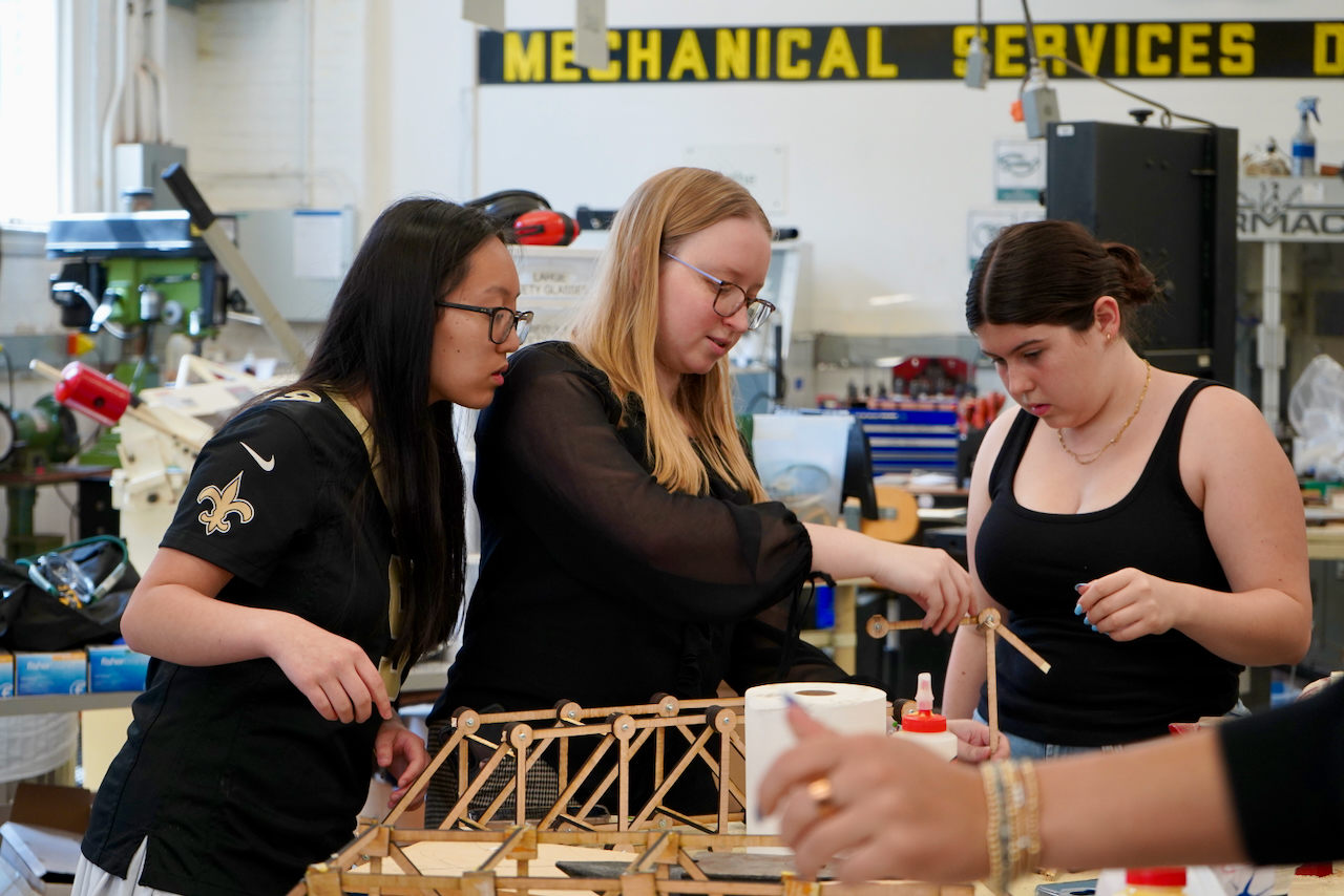 three girls looking at a wooden model of a bridge
