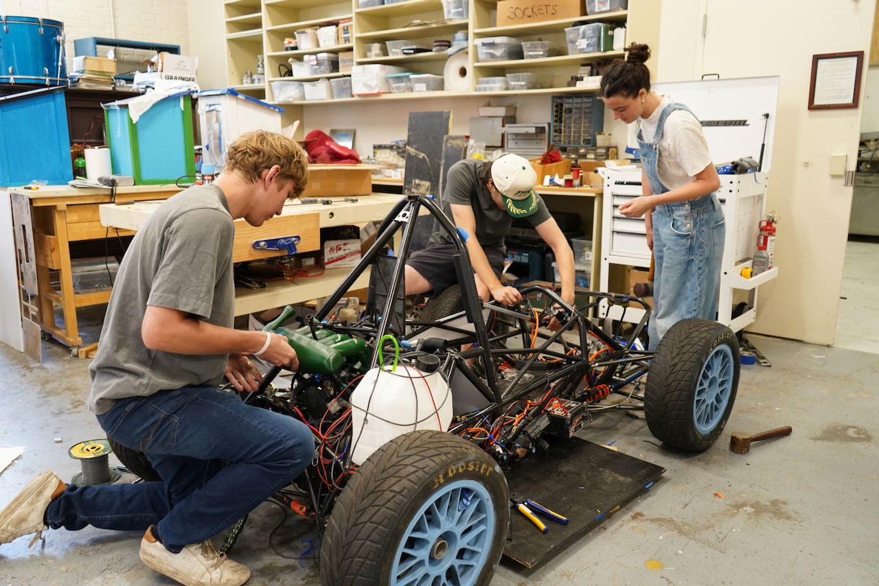 Three young people work on a race car in a workshop.