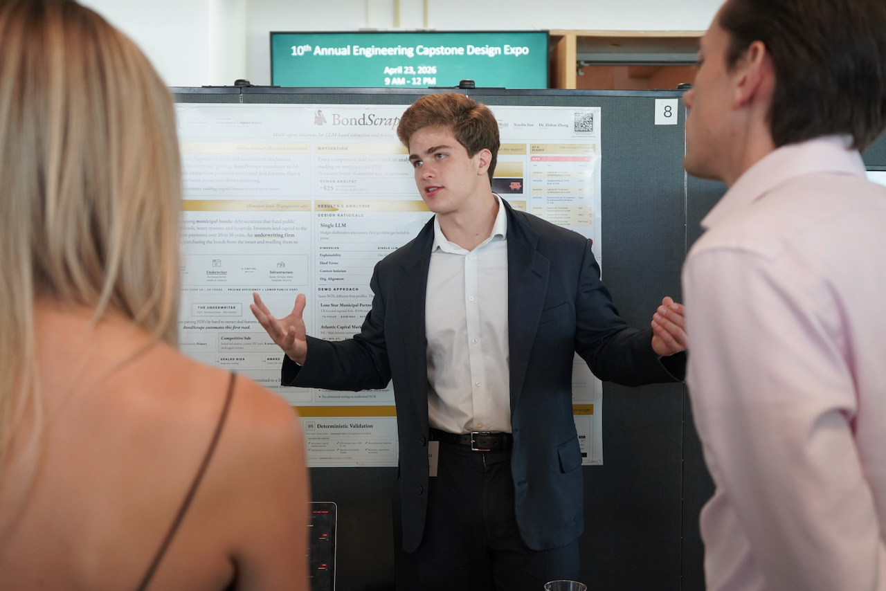 Photo of a young man in a suit presenting his project to two people.