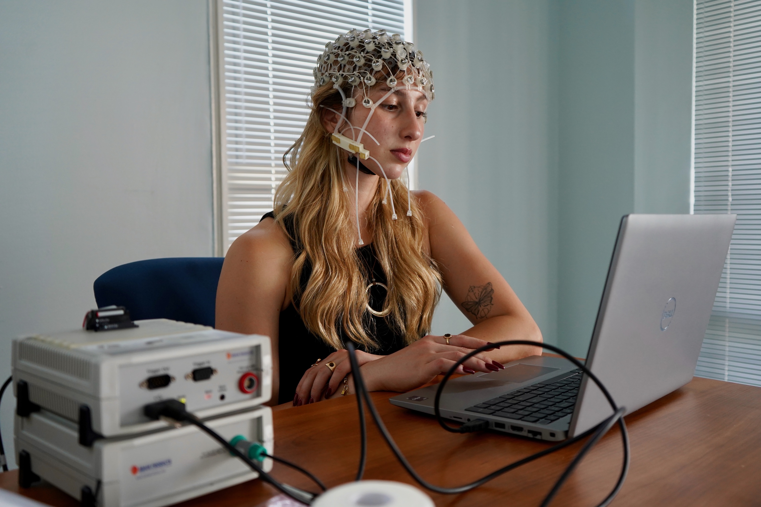 A woman wearing an EEG cap sits at a wooden desk with a laptop and testing equipment.