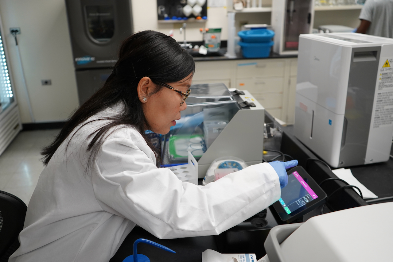 Photograph of a female scientist in a white coat pointing at a laboratory tablet.