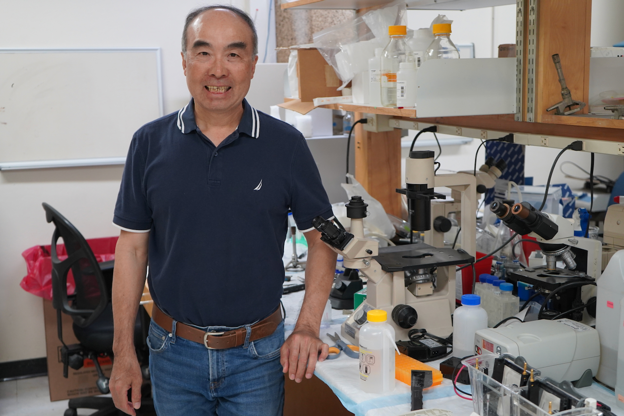 Photograph of an older Asian man standing in a laboratory, smiling at the camera.