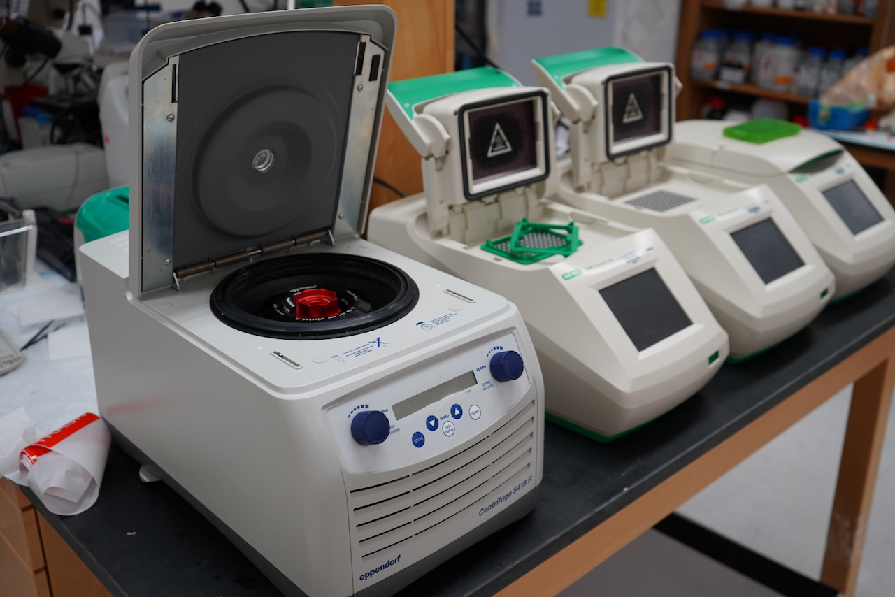Photograph of a centrifuge and three thermal cyclers lined up on a lab bench.