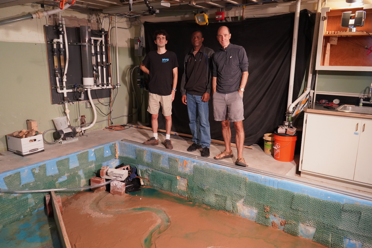 Three men stand behind a large, unfinished pool with a model river running through it.