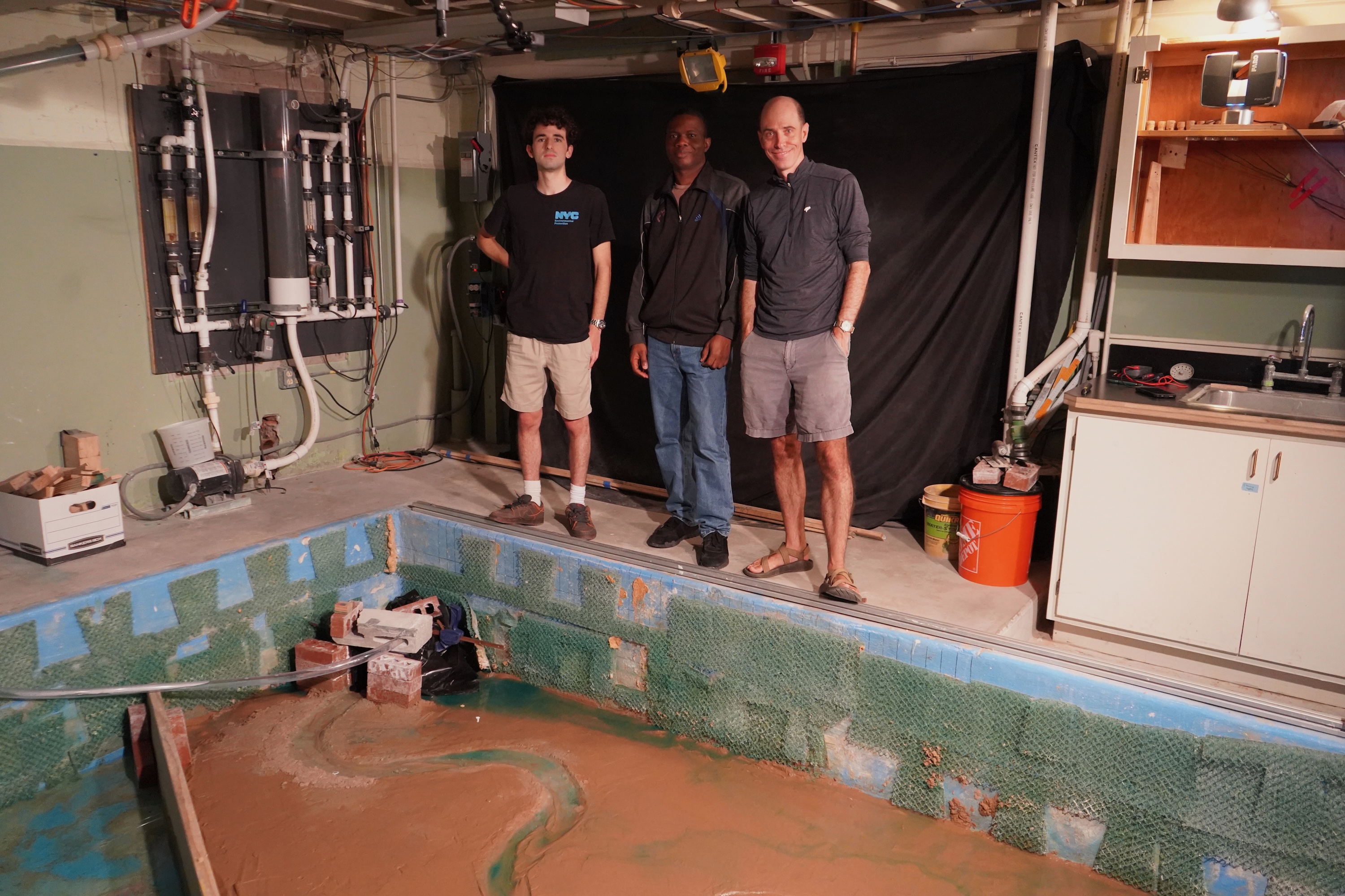 Three men pose by a large rectangular pool filled with muddy water in a basement.