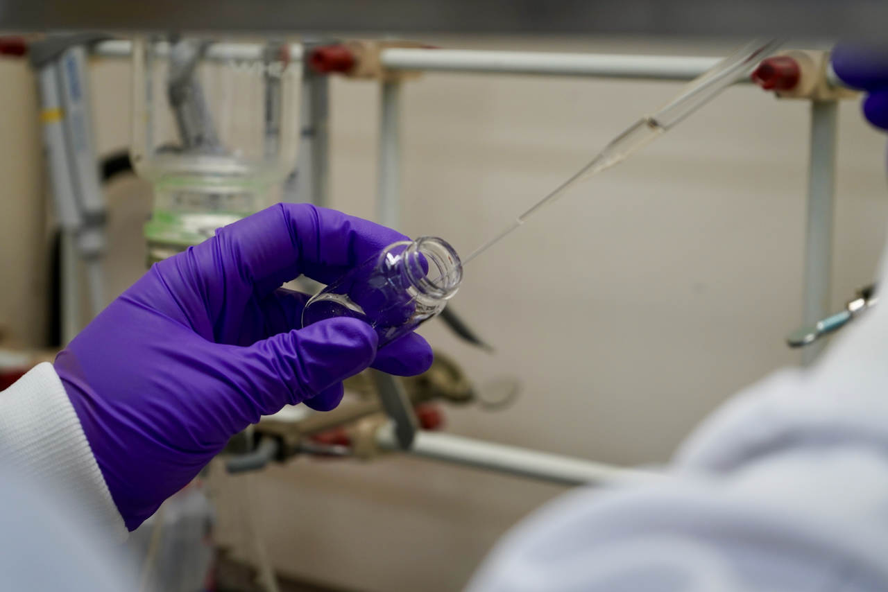 A gloved hand holds a glass vial while a pipette dispenses liquid into it in a lab.
